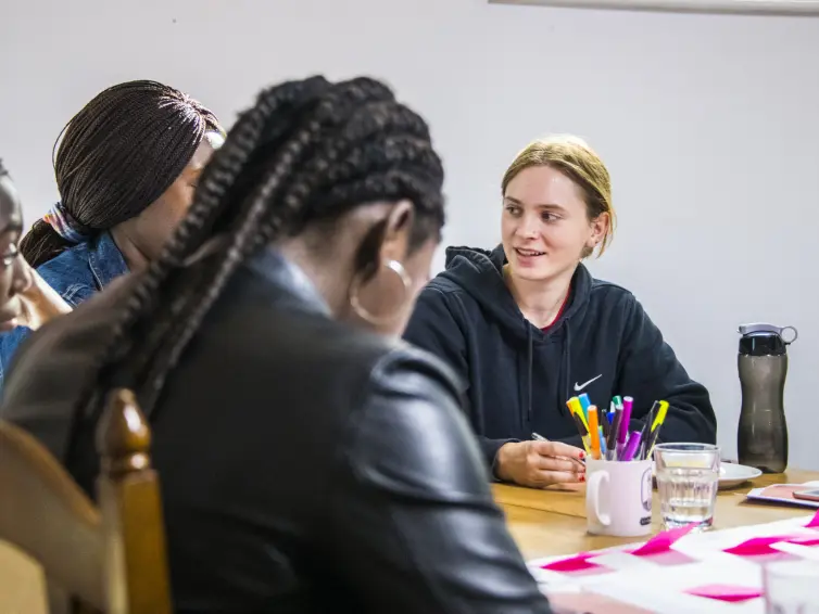 A young person smiling with other young people around a table. On the table there's a white page with bright pink post its on it and stationary scattered around.