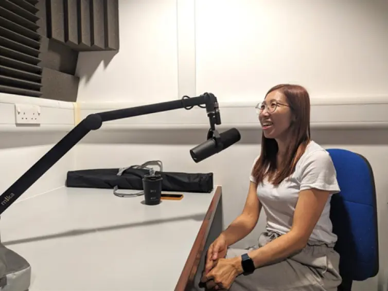 A person (Mon) at a desk with a microphone, smiling whilst talking.