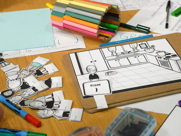 A tabletop shows cartoon character cutouts, colored markers, and a storyboard featuring a kitchen scene labeled 