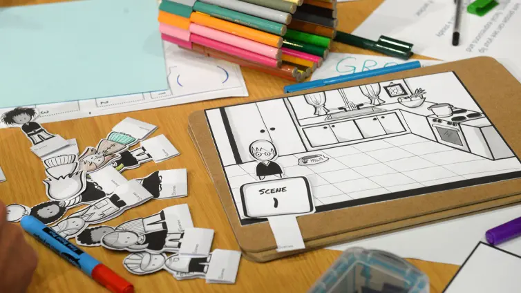 A tabletop shows cartoon character cutouts, colored markers, and a storyboard featuring a kitchen scene labeled 