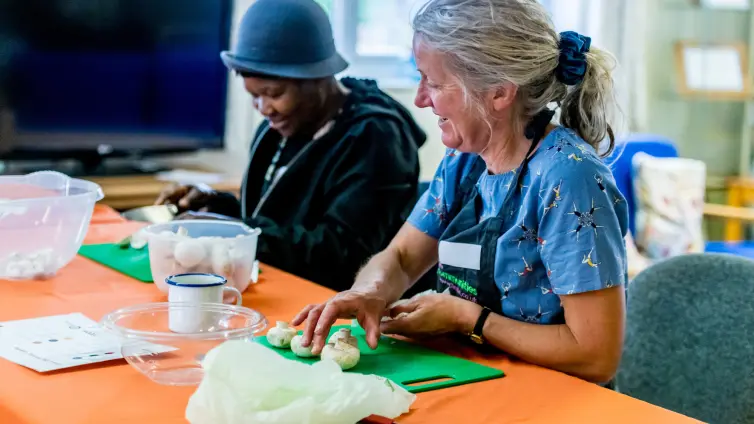 Two people chopping vegetables, one is smiling looking at the other one as she chops food.